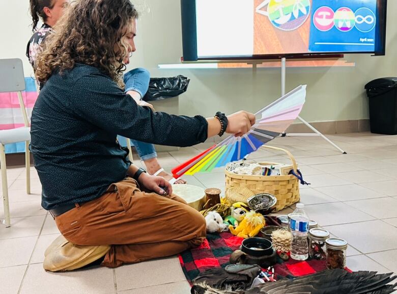 A young man with dark curly hair sits on the floor, waving a rainbow fan over smudging materials.