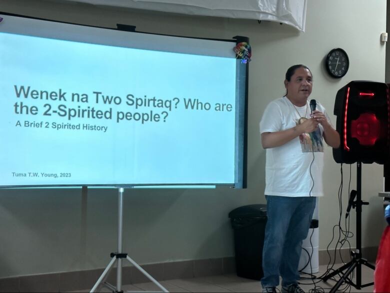 A man with dark hair and a white t-shirt holds a microphone. He stands in front of a power-point presentation that says Wenek na Two Spiritaq? Who are the 2-spirited people? 