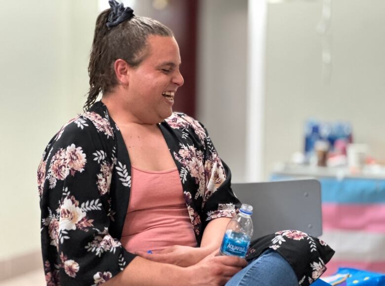 A person with a pony tail and pink shirt sits on a chair laughing. In the background is a blue, pink and white Trans flag.