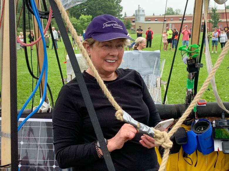 A woman with short blonde hair pulled back under a dark blue baseball hat and wearing a black long-sleeve shirt stands in the cockpit of a giant helium balloon aircraft holding a cell phone.