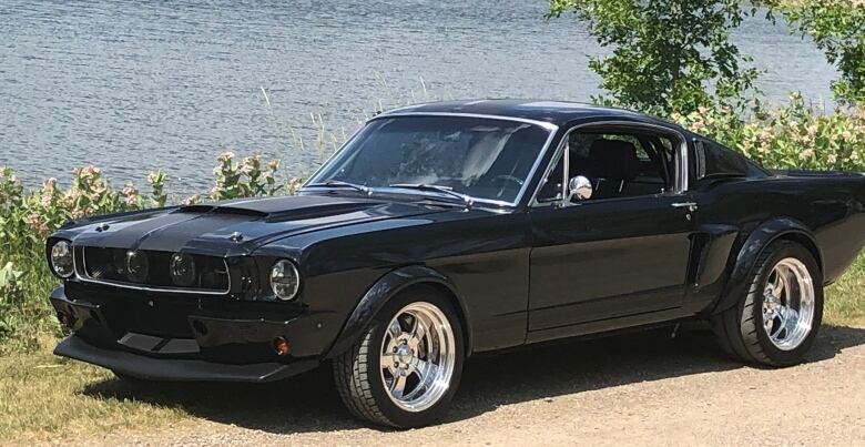 A black 1965 Ford Mustang coupe on a dirt rode next to a lake.