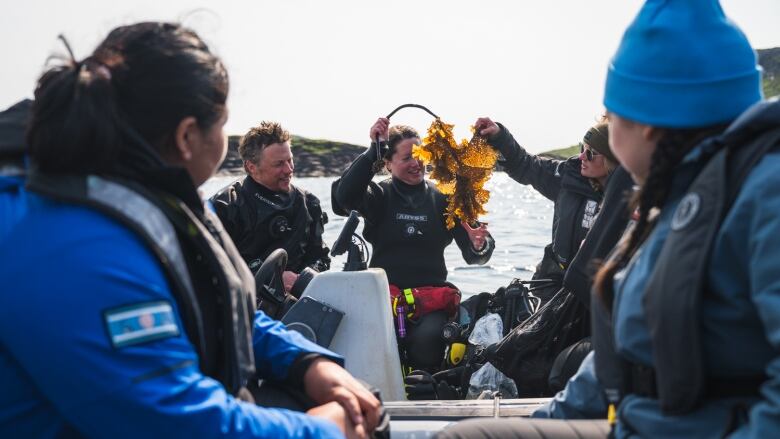 A woman holds up seaweed to a group of young people in a small boat. 