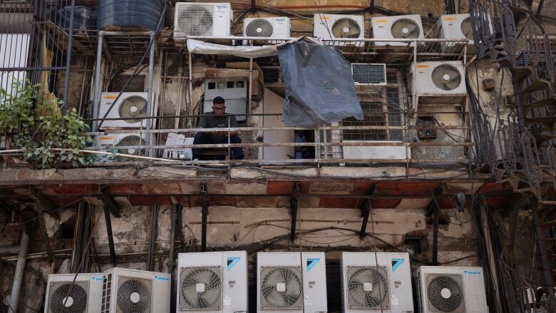 A man sits on the second floor of a multi-storey building, covered in air conditioners.