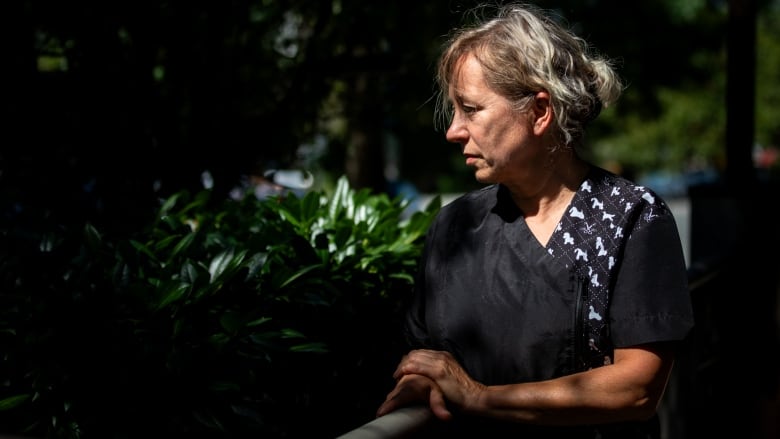 A blonde woman wearing a black dog-grooming smock stands in profile in partial shadow in front of a background of greenery.