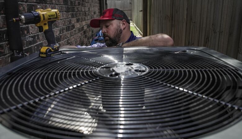 A man with a dark beard, wearing a red and black ball cap, fixes an outdoor air-conditioning unit.