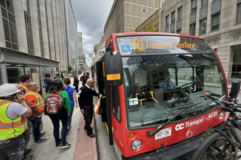 People board a red and white transit bus in a city's downtown in summer.