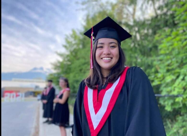 A teenage woman with black hair smiles while wearing graduation clothes.