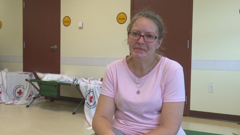 A woman in a pink t-shirt and glasses is sitting and talking to a reporter off-camera. A cot with a Red Cross blanket is in the background.