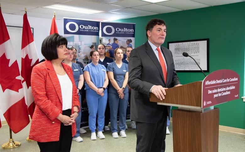 A woman and man stand in front of a podium making an announcement, with several people in nurses' uniforms in the background.