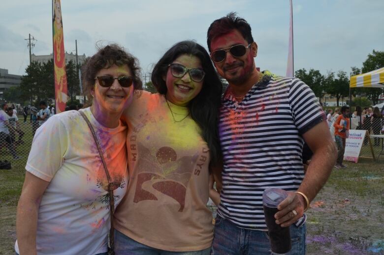 Three people stand next to each other covered in purple, yellow and orange powders after attending the colour throwing portion of the Festival of Colours.