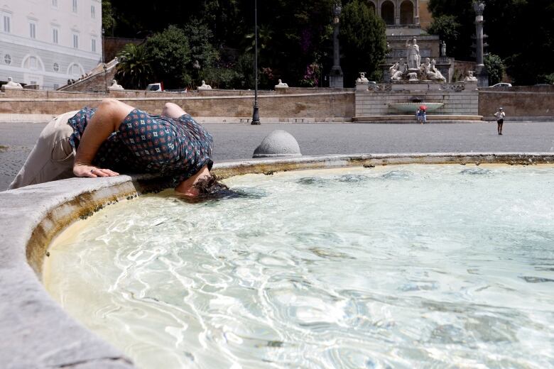 A person bends over to stick their head entirely underwater in a circular fountain. A statue is visible in the distance.