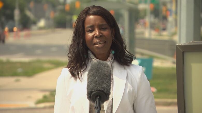 A woman wearing a white jacket speaks into a large fuzzy microphone outdoors.