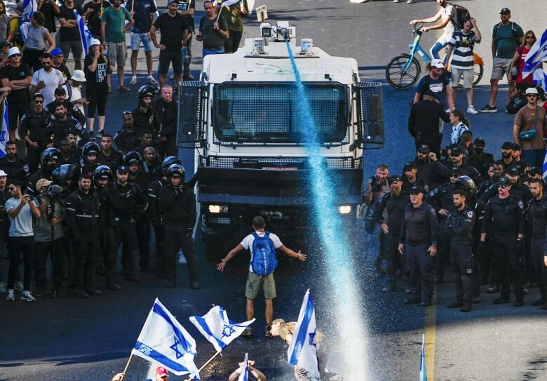 A police vehicle flanked by officers shoots liquid from a cannon at a group of protesters, some waving Israeli flags.