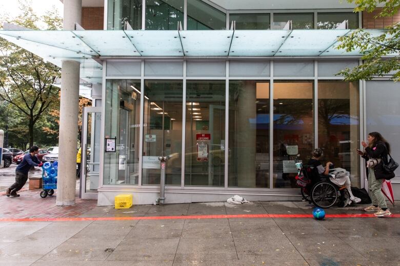 A few people mill around on a sidewalk next to a glass building.