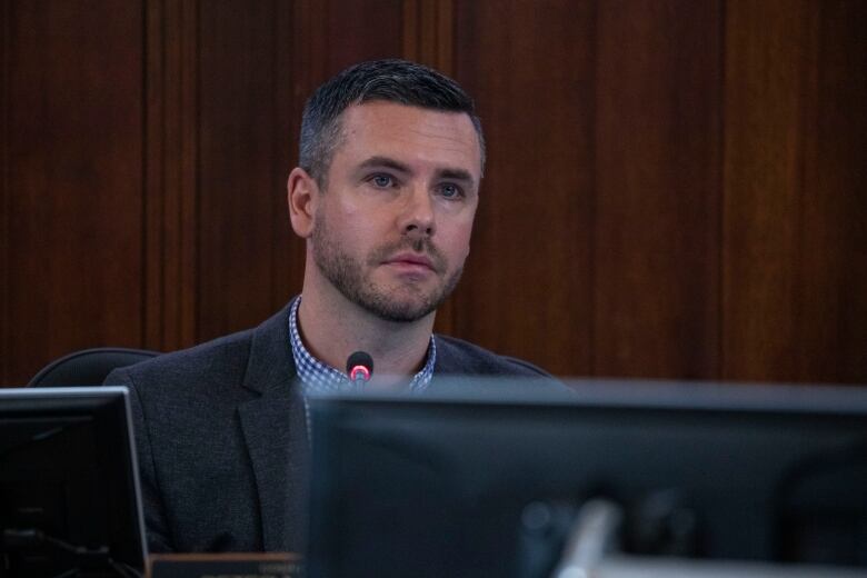 A man wearing a suit sits in council chambers with a mic in front of him.