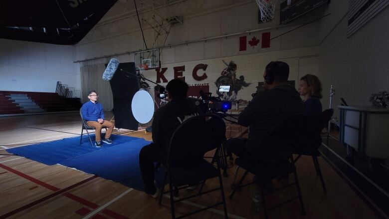 A man sits in a gymnasium as a film crew shoots an interview with him. 