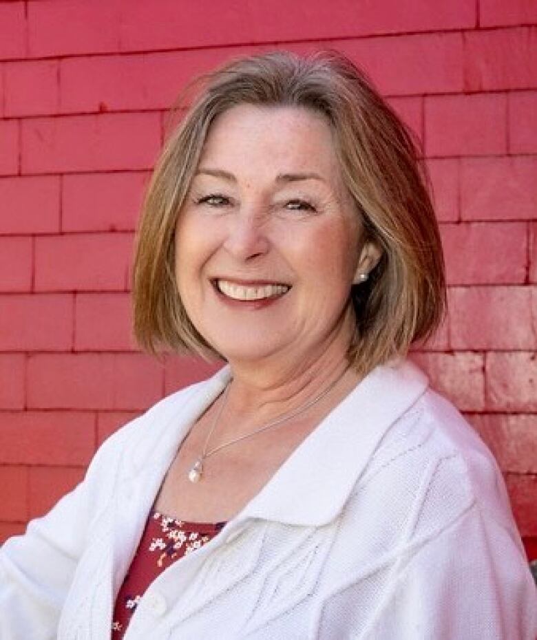 A white woman with shoulder-length dark blonde hair is wearing a long white shirt against the exterior red wooden-shingled wall of a house.