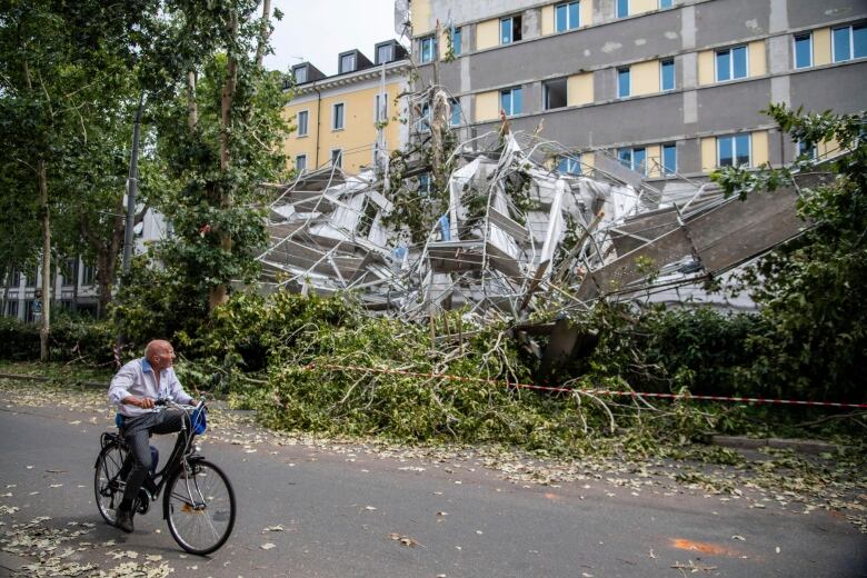 A bald man on a bicycle turns his head toward fallen trees and a crumple of metal that was presumably a structure.