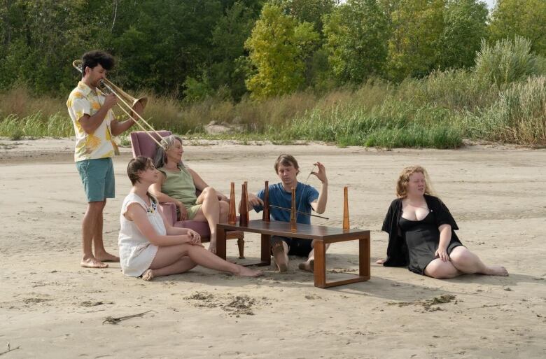 A person in a button down yellow shirt plays a trombone while other people sit around a table on a beach.