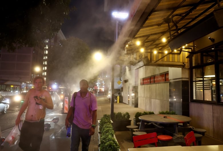 Two men, one with his shirt off, walk through a cloud of mist on a city sidewalk at night.