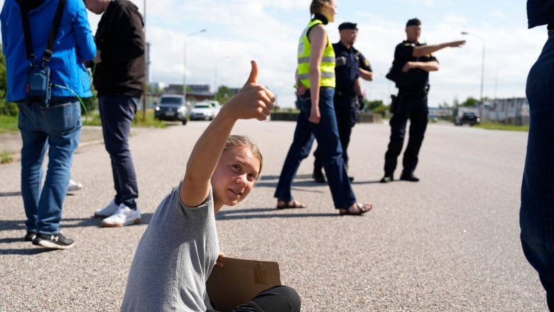 A young woman sitting cross-legged on a paved road looks behind her, making a thumbs-up gesture. Police and others mill about. 