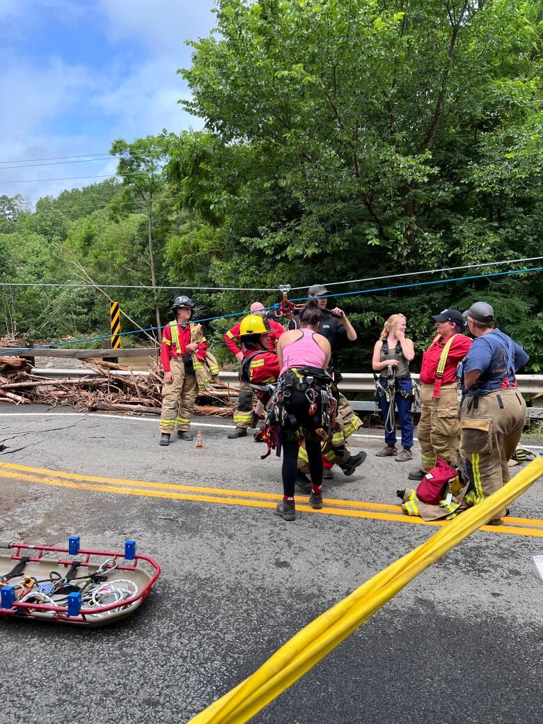 Several firefighters and people wearing ziplining harnesses stand on a road. There is a highline above them. 