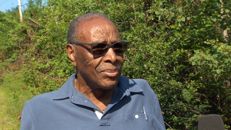 A black man wearing a short-sleeve shirt and sunglasses looks at the camera.