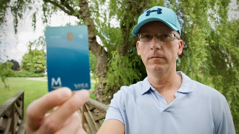 A man in a blue shirt holds up a blue credit card in a park.