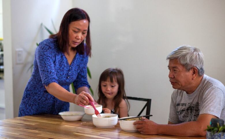 Two seniors sit at a table with a young girls looking at bowls of noodles. The young girl has her tongue sticking slightly out of her mouth with an eager look on her face. 