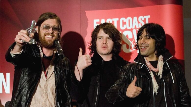 Three men stand in front of a red wall that says 