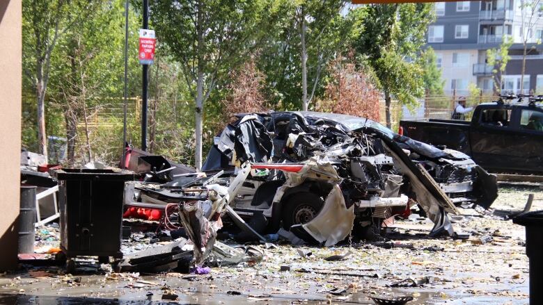 A truck is a mangled wreck after an explosion in a parking lot nearly obliterated it. There are pieces of metal and debris littered across the ground.