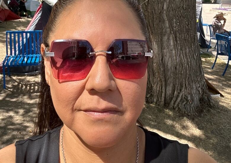 A woman with red sunglasses with her hair pulled back into a ponytail takes a selfie with a serious face.