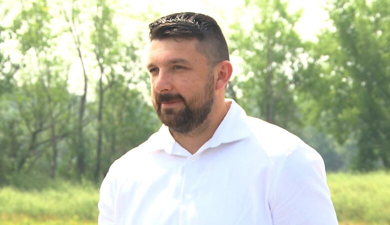 A man with a crew cut and a white shirt speaks to the camera in a natural environment.