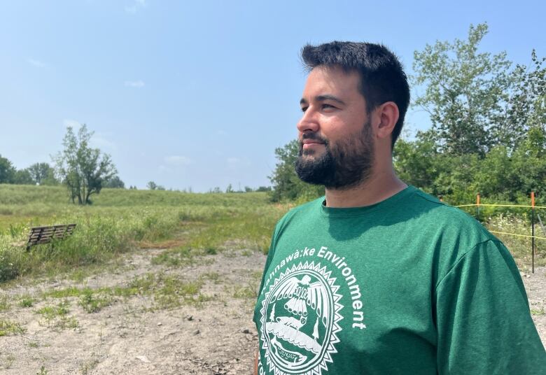 A man wearing a green shirt that says Kahnaw:ke Environment on it looks out onto a marshy bay.