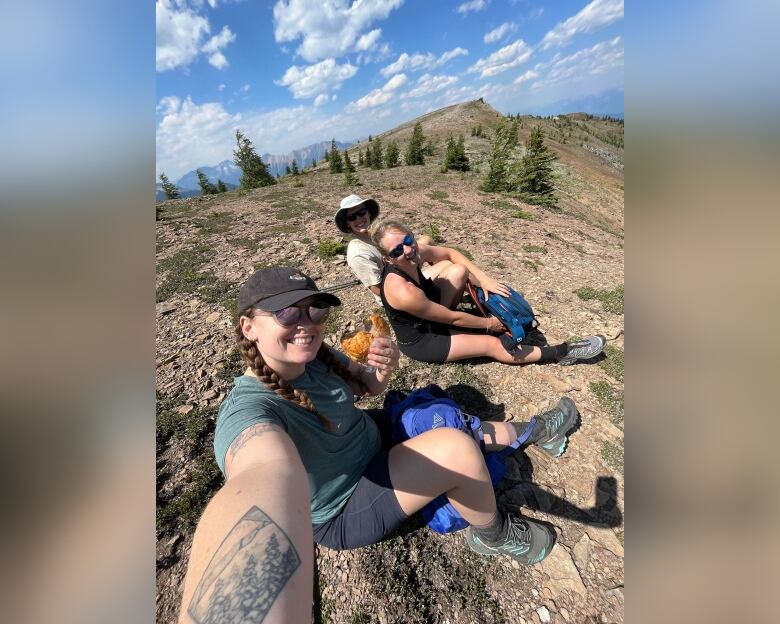 Three people sit on a mountain ground, with the left one holding a sandwich.