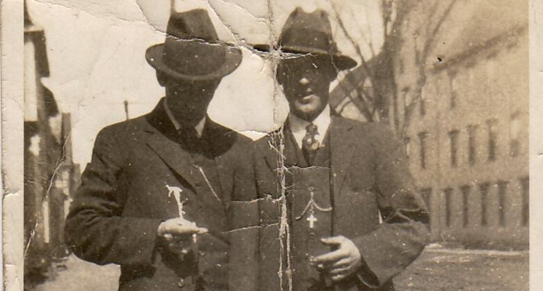 Two men in a sepia toned photo. They stand in front of an old building wearing suits and fedoras. 