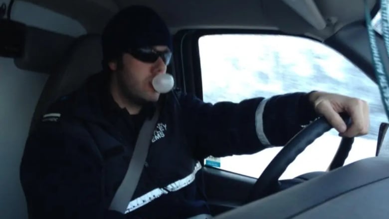 A man in an EMS uniform drives an ambulance and is blowing a bubble with gum. 