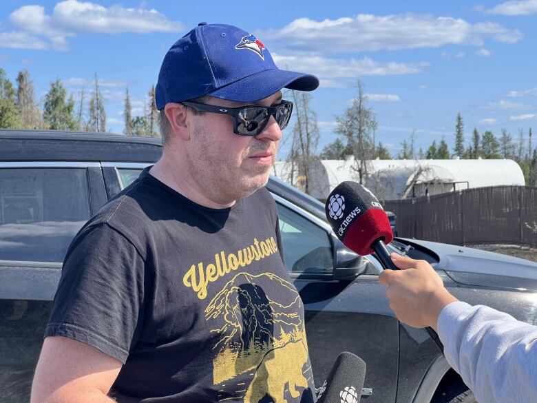 A man in sunglasses and a ballcap stands beside a truck as someone's arm holds a CBC microphone in front of him.