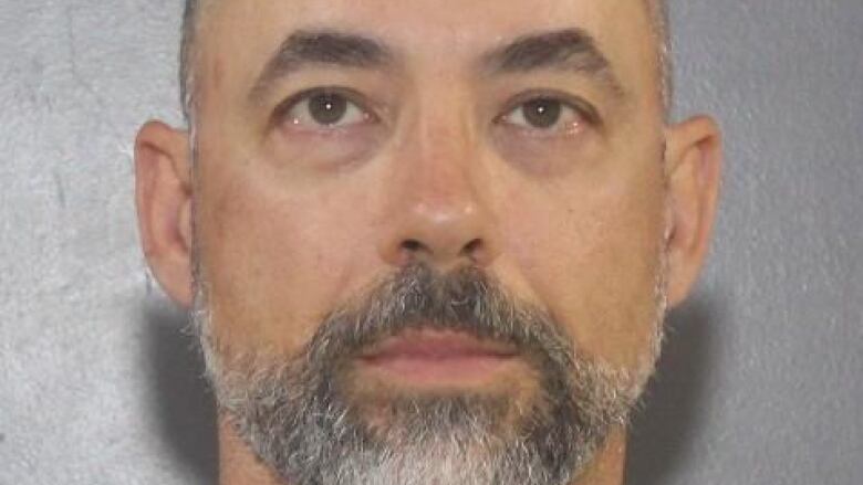 A mug shot shows a bald white man with a short grey beard in front of a grey backdrop.