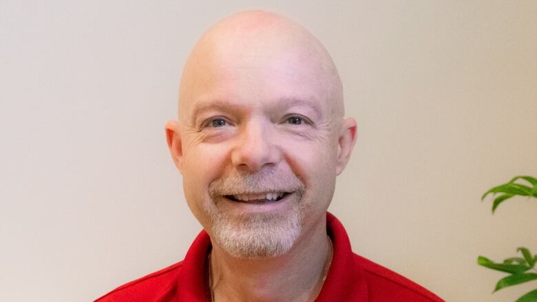 A smiling white man with a short white beard is shown wearing a red polo shirt in front of a beige background.