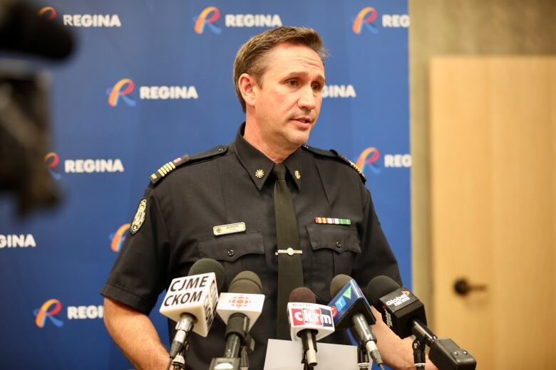 A man in a black uniform speaks at a podium featuring a variety of media microphones. 