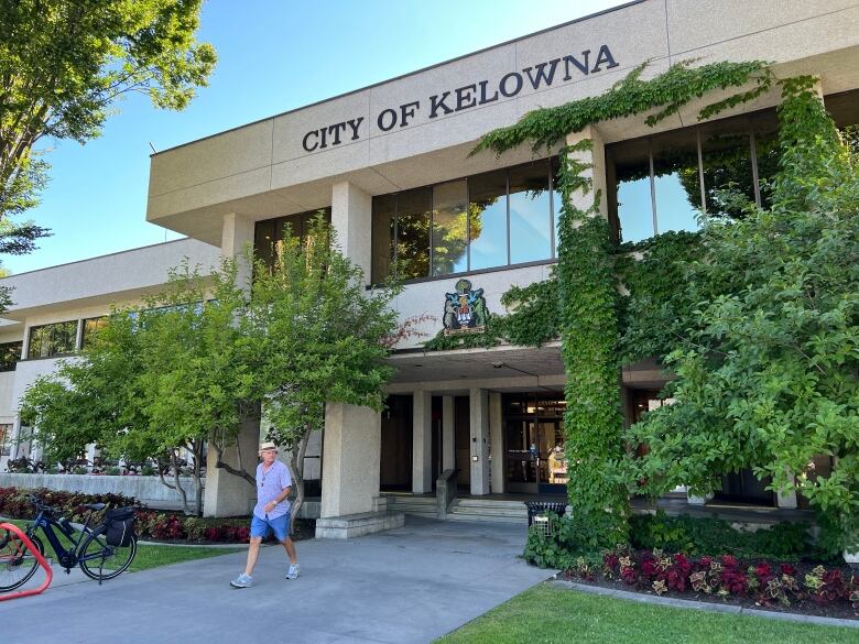 A man walks by a building with ivy on its pillars and the name 'City of Kelowna' over the entrance on a sunny day. The building is bordered by a flower bed with red pansies and a dark green lawn.