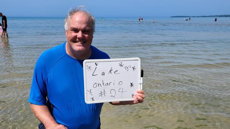 David kneels in the water at a beach with a small white board in his hand. The white board reads, 