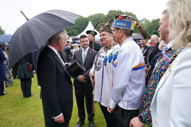 A person holding an umbrella and top hat speaks with other people wearing Indigenous dress.