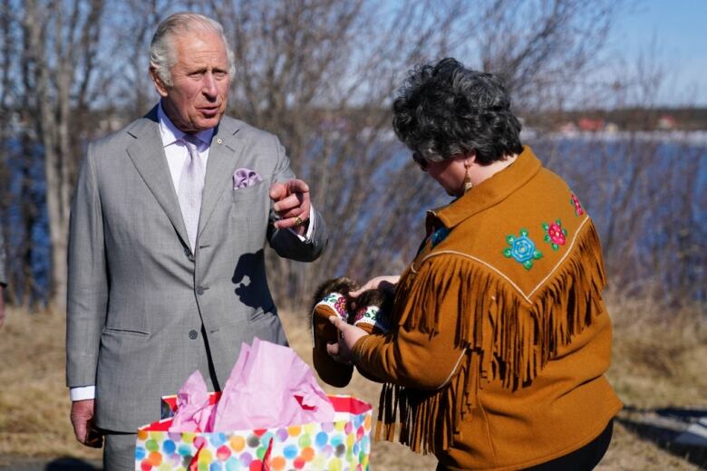 A person talks to another person holding moccasins near a riverbank.