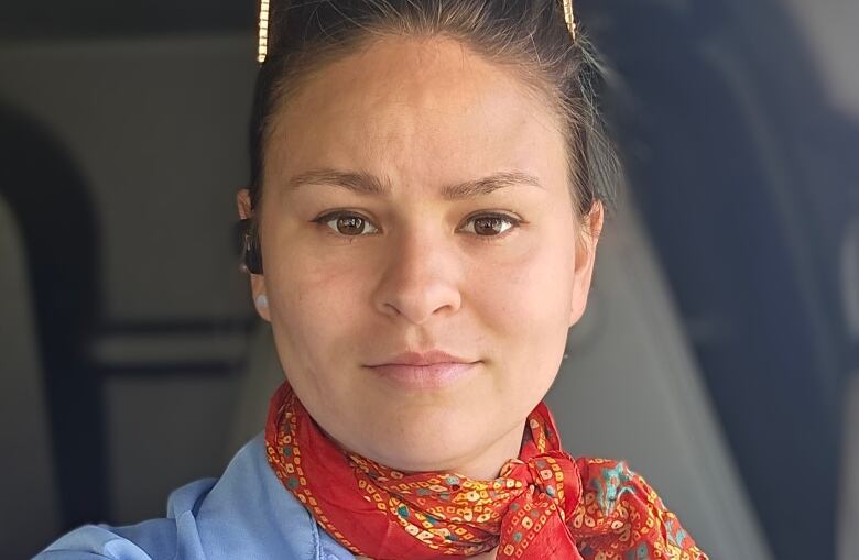 A woman with blonde hair pulled back looks straight into the camera. The woman is wearing a blue shirt with a red scarf around her neck.