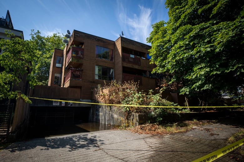 Caution tape is slung across the entrance to a lowrise brownstone apartment building surrounded by trees.