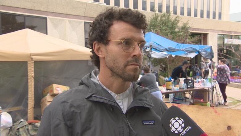 A white man white brown hair and trim beard is wearing glasses and a charcoal grey coat over a white shirt. Behind him are tents and boxes, and Regina's city hall. Some volunteers are handing out items at one of the tents.