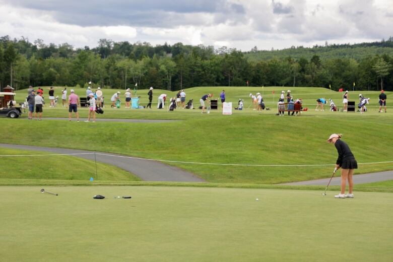 One golfer practices a long put on a putting green while others practice on the driving range behind her. 