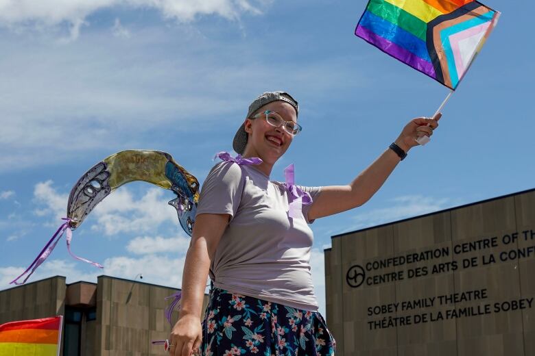 Woman wearing a ball cap backwards, glasses, a gray t-shirt and flower-patterned skirt, waving a Pride flag. She is also sporting a pair of wings. She's in a Pride parade and going by a building that says, 